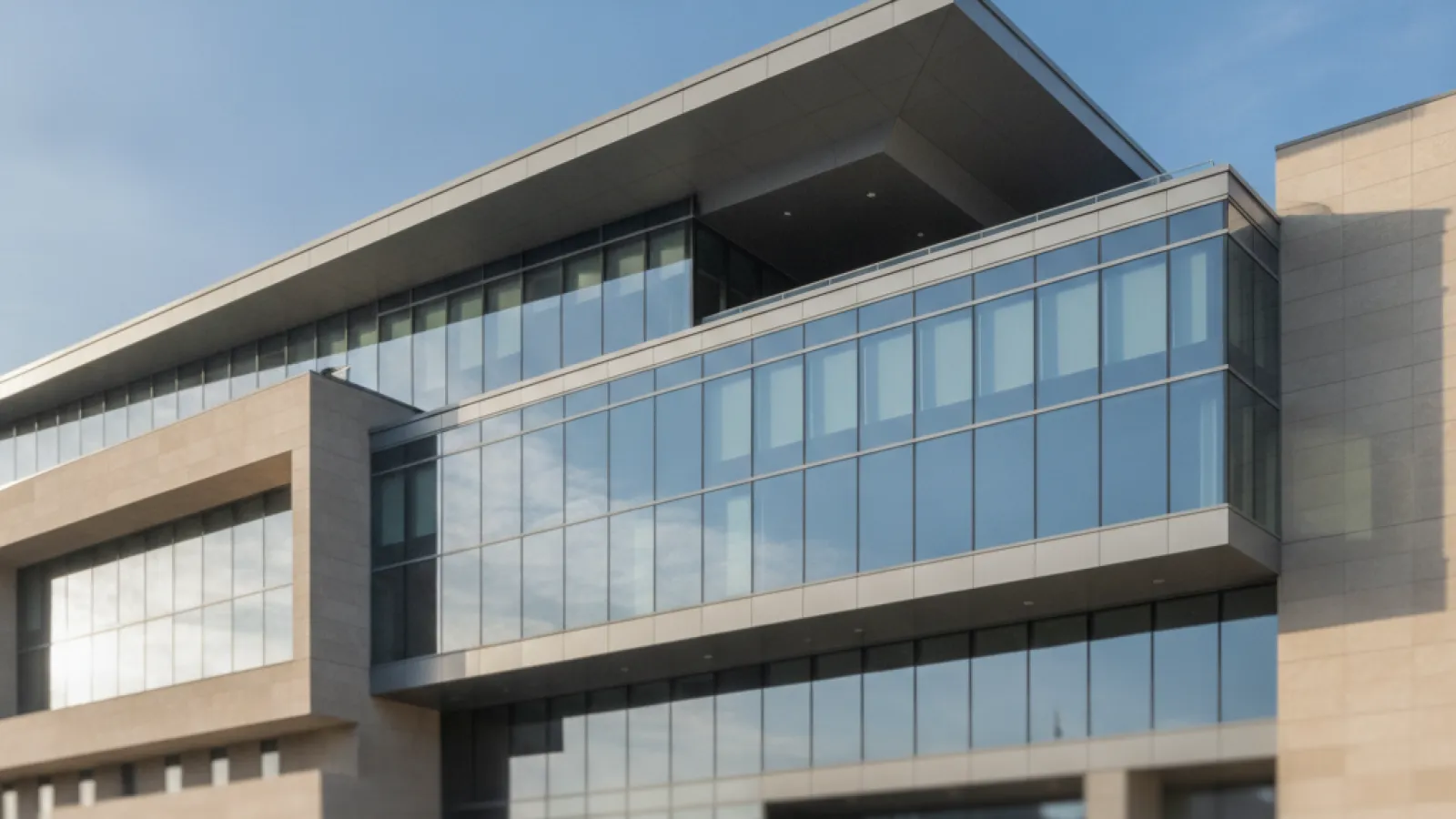 An exterior view of a modern building featuring a large glass facade, light stone sections, a wide overhanging roof, and a prominent elevated glass-enclosed section, illuminated by bright daylight under a clear blue sky.