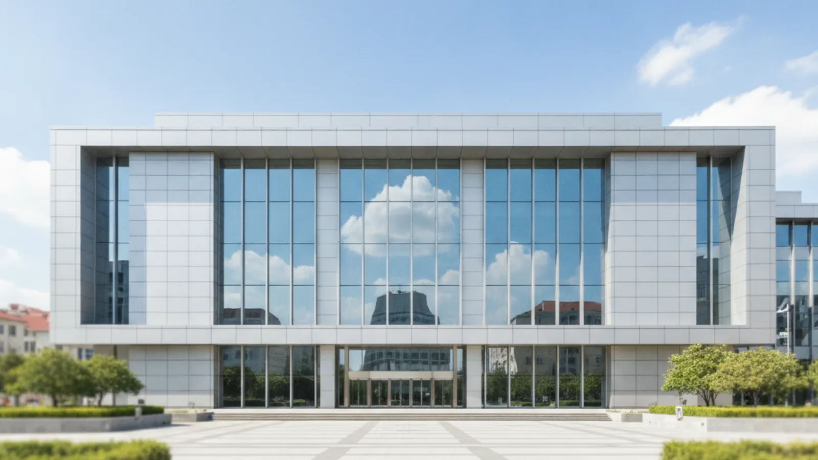 A modern building with a facade of large glass windows reflecting the sky and clouds stands behind a wide, patterned paved plaza on a clear, sunny day.